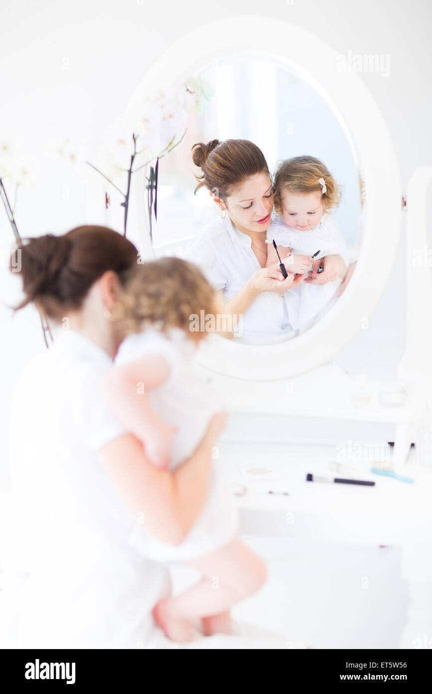 Junge attraktive Frau und ihre niedlichen Kleinkind Tochter Anwendung Make-up und Kosmetik mit Bürste sitzt vor der Runde Spiegel Stockfoto