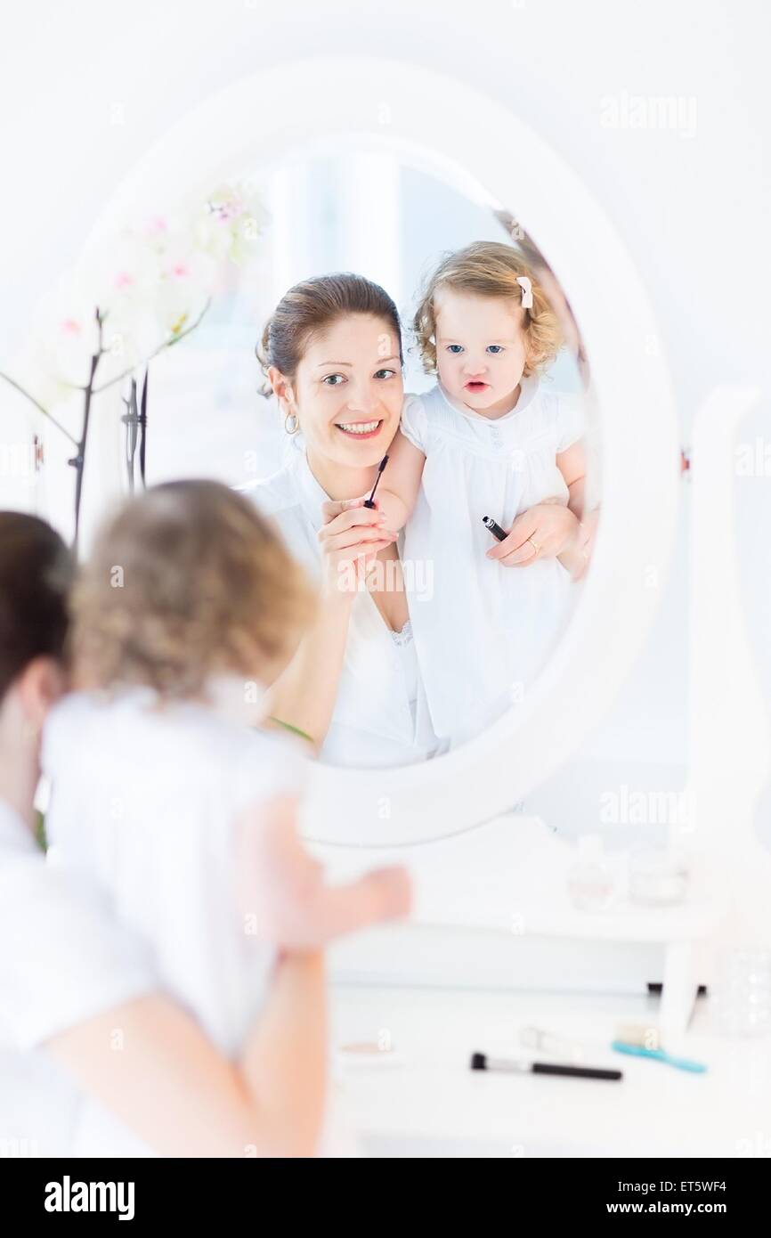 Junge attraktive Frau und ihre niedlichen Kleinkind Tochter Anwendung Make-up und Kosmetik mit Bürste sitzt vor der Runde Spiegel Stockfoto