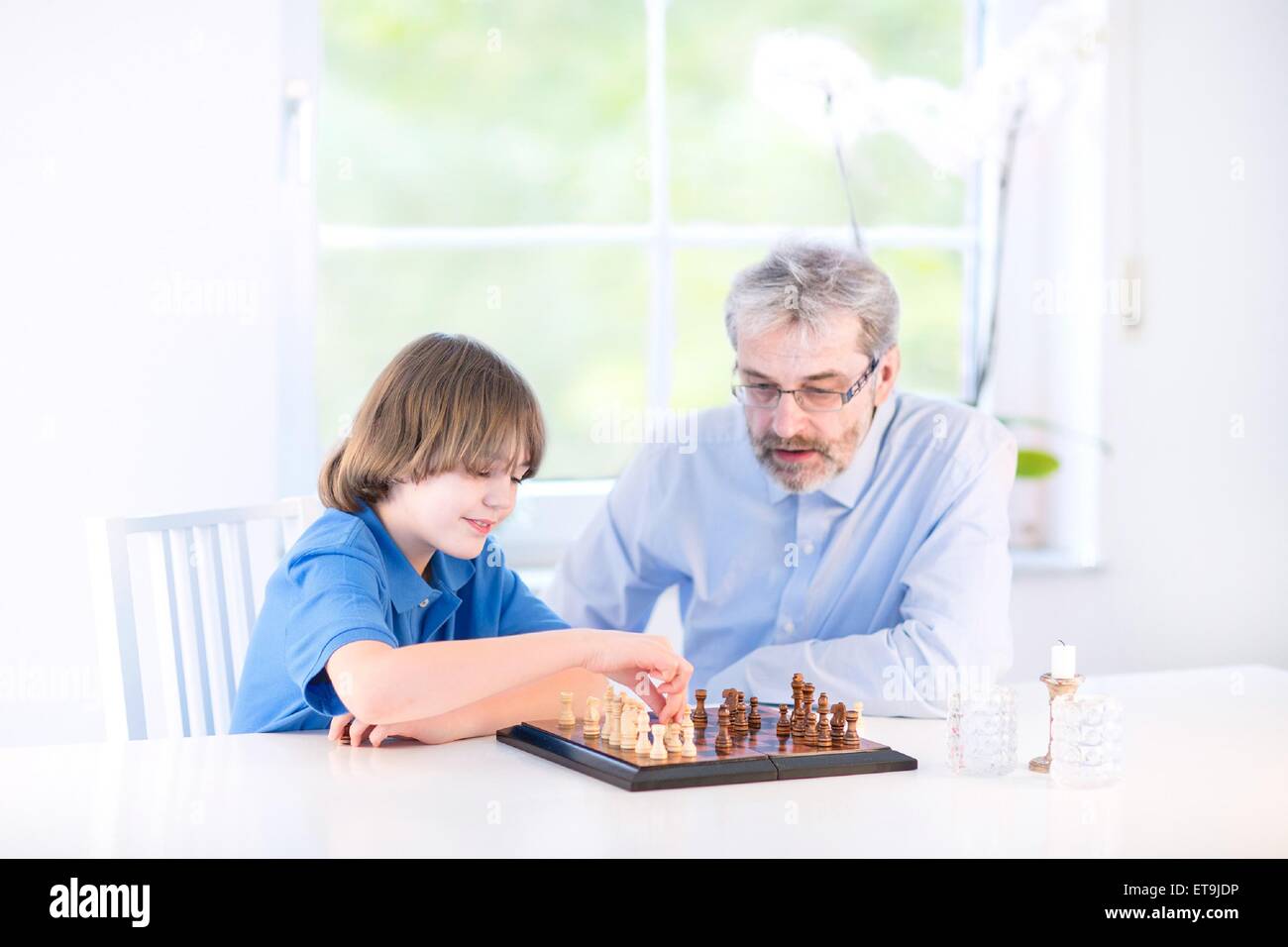 Niedlich fröhlicher Junge spielt Schach mit seinem Großvater sitzt am Fenster in einem weißen Wohnzimmer Stockfoto