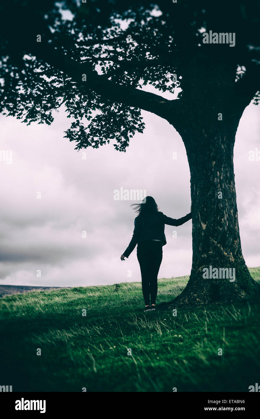 Silhouette einer weiblichen Figur lehnt sich an einen Baum in der Landschaft Stockfoto