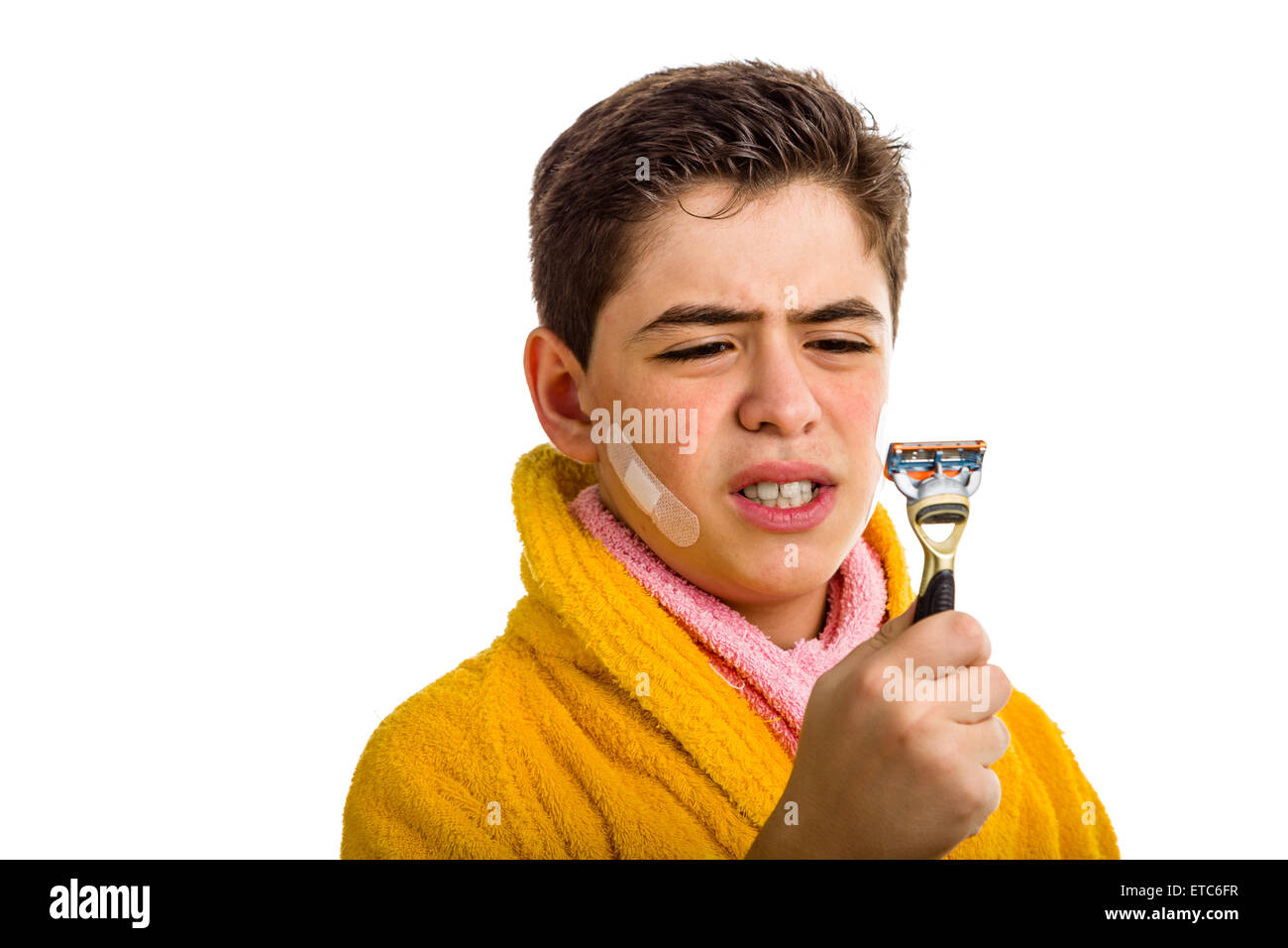 Ein spanischer Junge trägt einen gelben Bademantel mit einem rosa Handtuch um den Hals: er hat einige Flecken auf seinem Gesicht und starrt wütend auf das Rasiermesser er für die Rasur verwendet Stockfoto