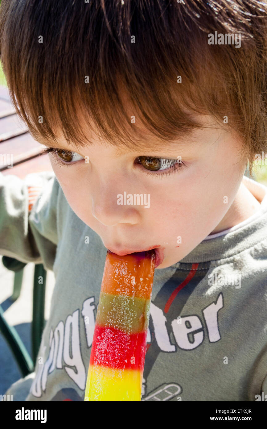 Oben nah am Gesicht des Kindes, Junge, 4-5 Jahre alt, beißt sich in einen mehrfarbigen Eislastwagen, während es im Freien bei Sonnenlicht liegt. Stockfoto