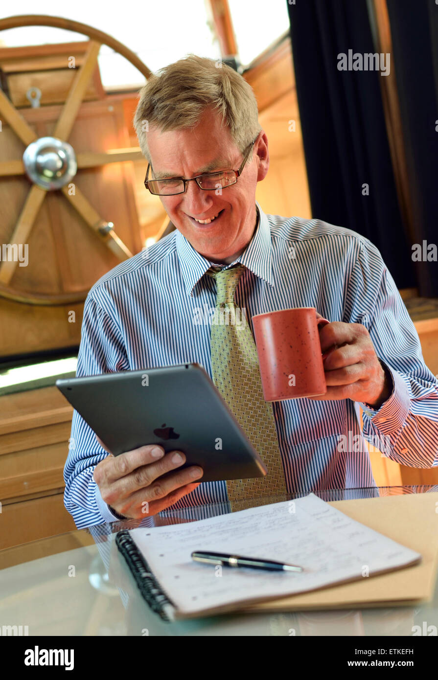 IPad Business man Online-Zoom-Teams Treffen reifen Unternehmer mit Tee Interaktion mit seinem iPad Air smart Tablet-Computer im Binnenschiff Hausboot Büro Stockfoto