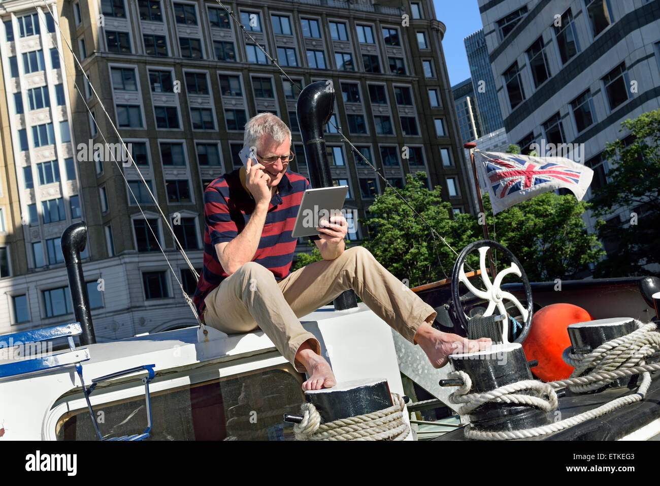 CANARY WHARF reifer Geschäftsmann sitzt draußen auf seinem Büroboot und spricht auf seinem Smartphone und schaut auf sein iPad, Büros hinter Canary Wharf London E14 Stockfoto