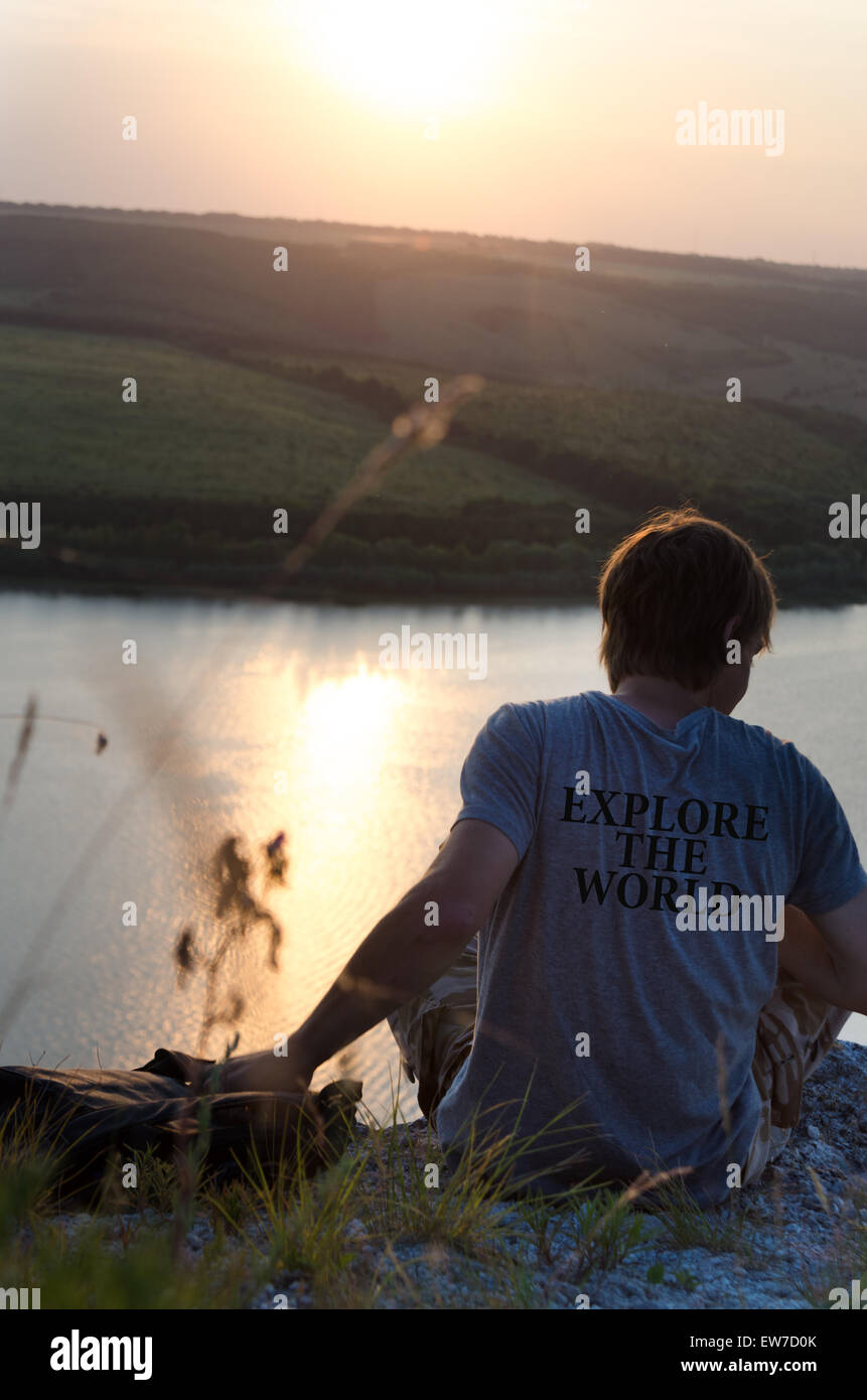 Mann sitzt auf einem Felsen über dem Fluss bei Sonnenuntergang, Reise-Konzept. Entdecken Sie die Welt - auf dem T-shirt geschrieben Stockfoto