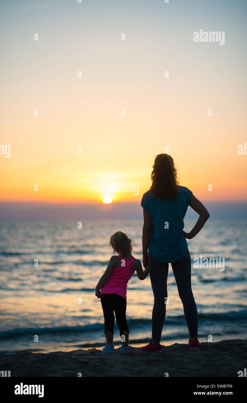 Mutter und junge Tochter halten Sie Hände, im Hintergrund schauen, wie die Sonne ins Meer am Horizont verschwindet. Sonnenuntergang am b Stockfoto
