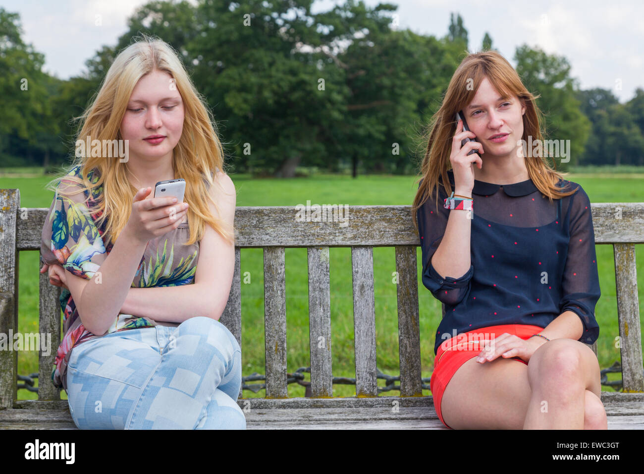 Zwei kaukasischen Teenager Freundinnen sitzen auf Holzbank im Park telefonieren mit Handy Stockfoto