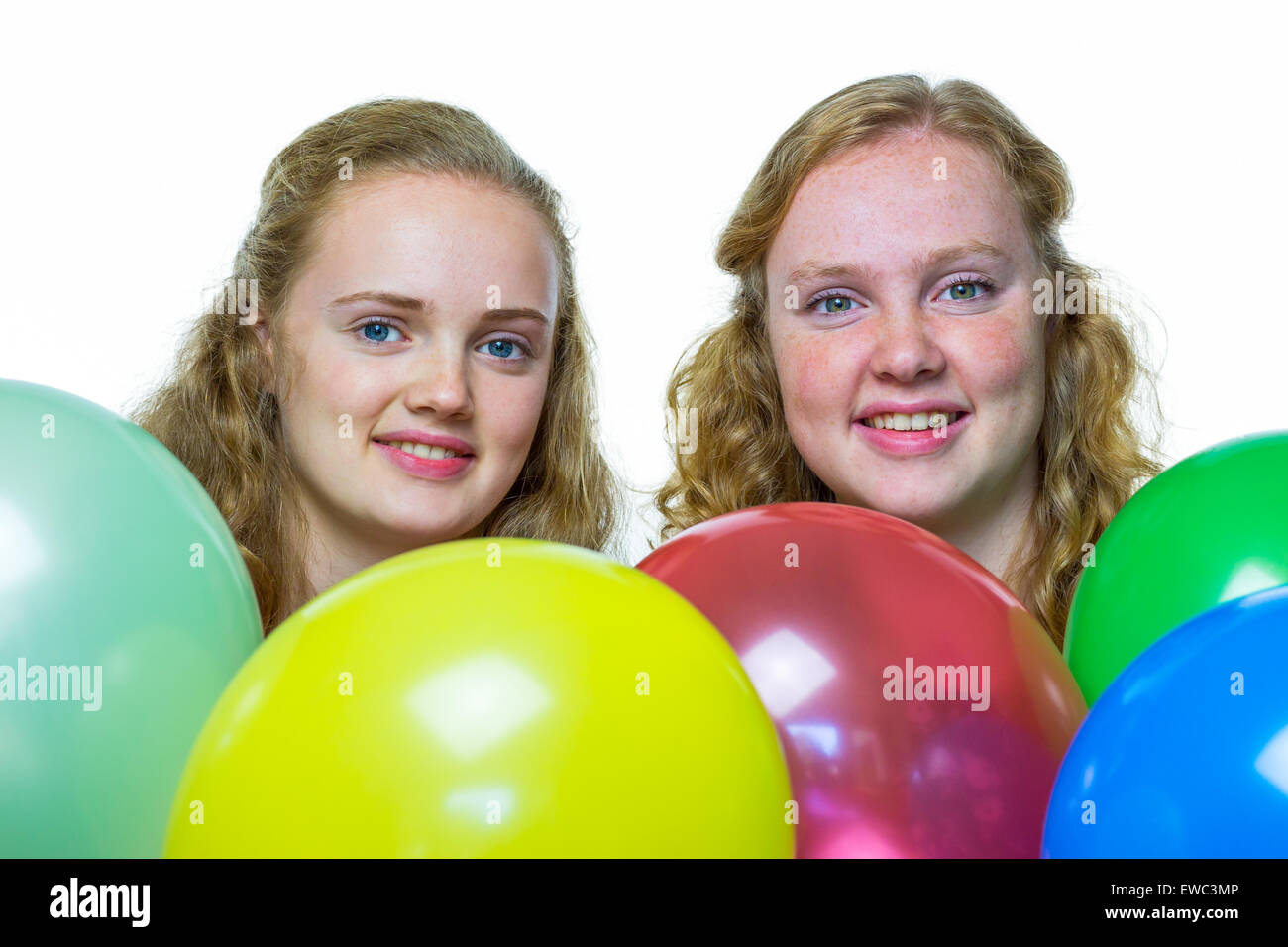 Zwei lächelnde kaukasische Mädchen im Teenageralter hinter verschiedenen bunten Luftballons, die isoliert auf weißem Hintergrund Stockfoto