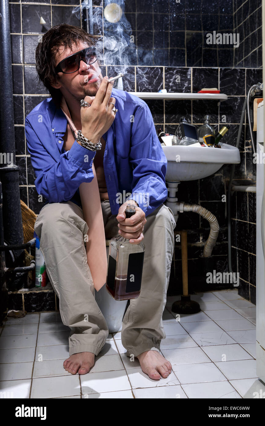 Betrunkener Mann sitzt in einer Toilette mit einer Flasche Whisky und Rauchen, Farbbild Stockfoto