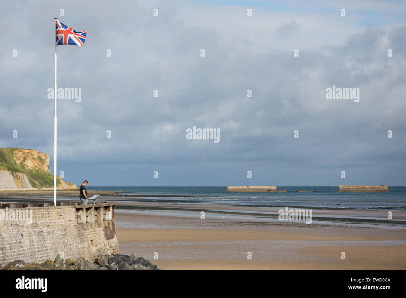 Ein einsamer Mann mit Kamera lesen eine Informationstafel über Arromanches Beach, Normandie, Frankreich, mit Anschluß-Markierungsfahne fliegen Stockfoto