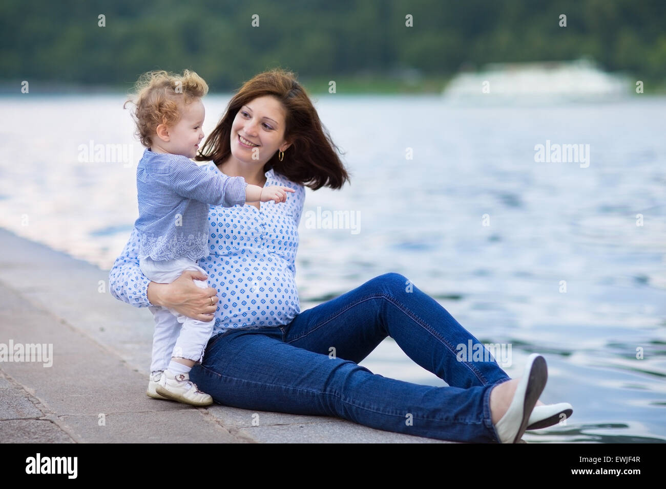 Junge schwangere Mutter und ihre kleine Baby-Tochter am Ufer eines Flusses in einem Stadtzentrum entspannend Stockfoto