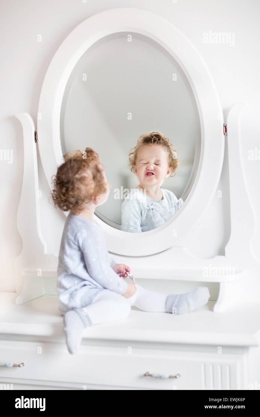 Sehr lustige Babymädchen mit dem lockigen Haar Blick auf ihr Spiegelbild in ein schönes weißes Schlafzimmer mit einer klassischen Kommode mit einem runden Stockfoto