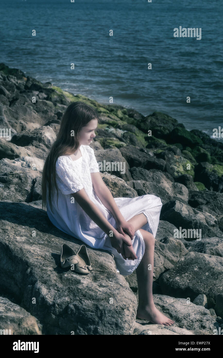 eine Mädchen sitzt barfuß auf einem Felsen am Meer, ihre Schuhe neben ihr Stockfoto