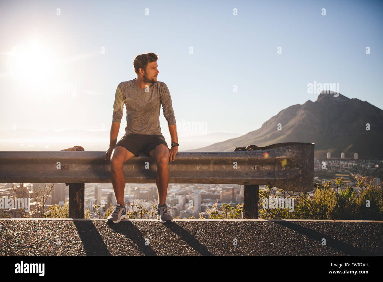 Männliche Läufer sitzt auf einem Geländer auf Land Straße wegsehen an sonnigen Tag. Junger Mann eine Pause nach dem Morgenlauf im freien Stockfoto