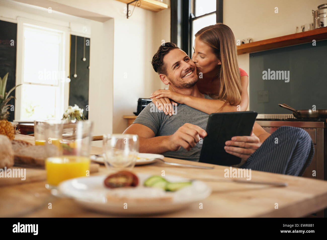 Junger Mann hält eine digitale-Tablette während seiner Freundin umarmt ihn von hinten, was ihm einen guten Morgen Kuss. Junge Liebespaar Stockfoto