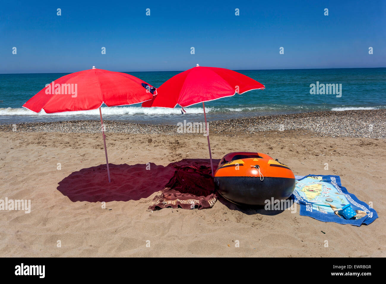 Zwei rote Sonnenschirme am Strand von Rethymno, Kreta, Griechenland Sonnenschirme werfen einen Schatten, Stillleben am Strand Aufblasbares Boot und Handtücher, niemand Stockfoto
