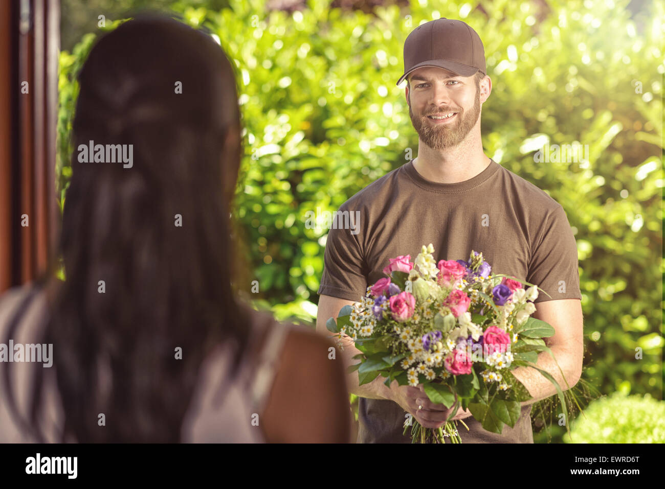 Lächelnde bärtigen 20er Jahre Mann tragen braune Haube und braunen T-shirt liefert Blumen an Tür der weibliche junge Brünette. Über die Schulter Stockfoto