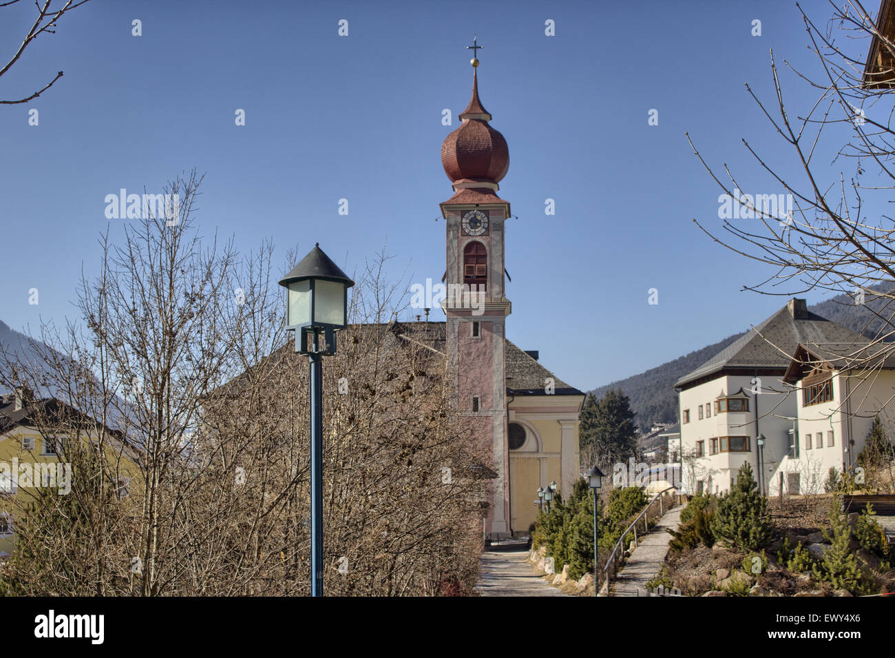 Der Luis Trenker Promenade führt bis St. Ulrich, italienisches Dorf in Dolomiten Alpen: typische Häuser, Gebäude und die roten Glockenturm mit Uhr Pfarrkirche St. Ulrich mit schneebedeckten Bergen und grünen Koniferen im Hintergrund Stockfoto