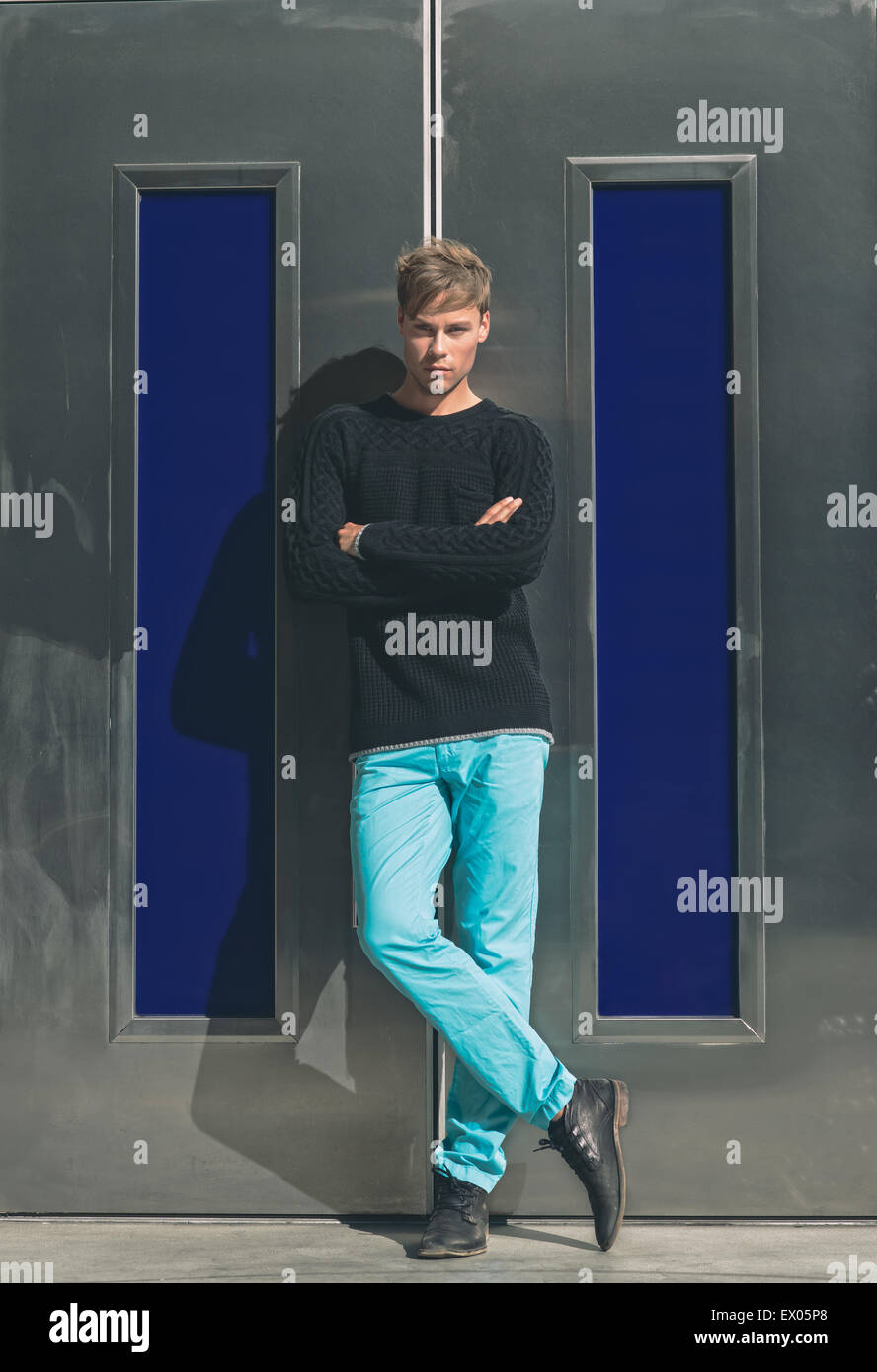 Ein stattlicher Mann, männliches Modell posiert im Freien gegen einen modernen, silbernen Tür mit seine Arme und Beine gekreuzt, ein Winter Männermode redaktionelle Konzept. Stockfoto