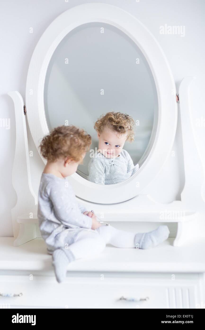 Lustige und niedliche Babymädchen beobachten ihr Spiegelbild in einem weißen Schlafzimmer mit einen schönen runden Spiegel Stockfoto