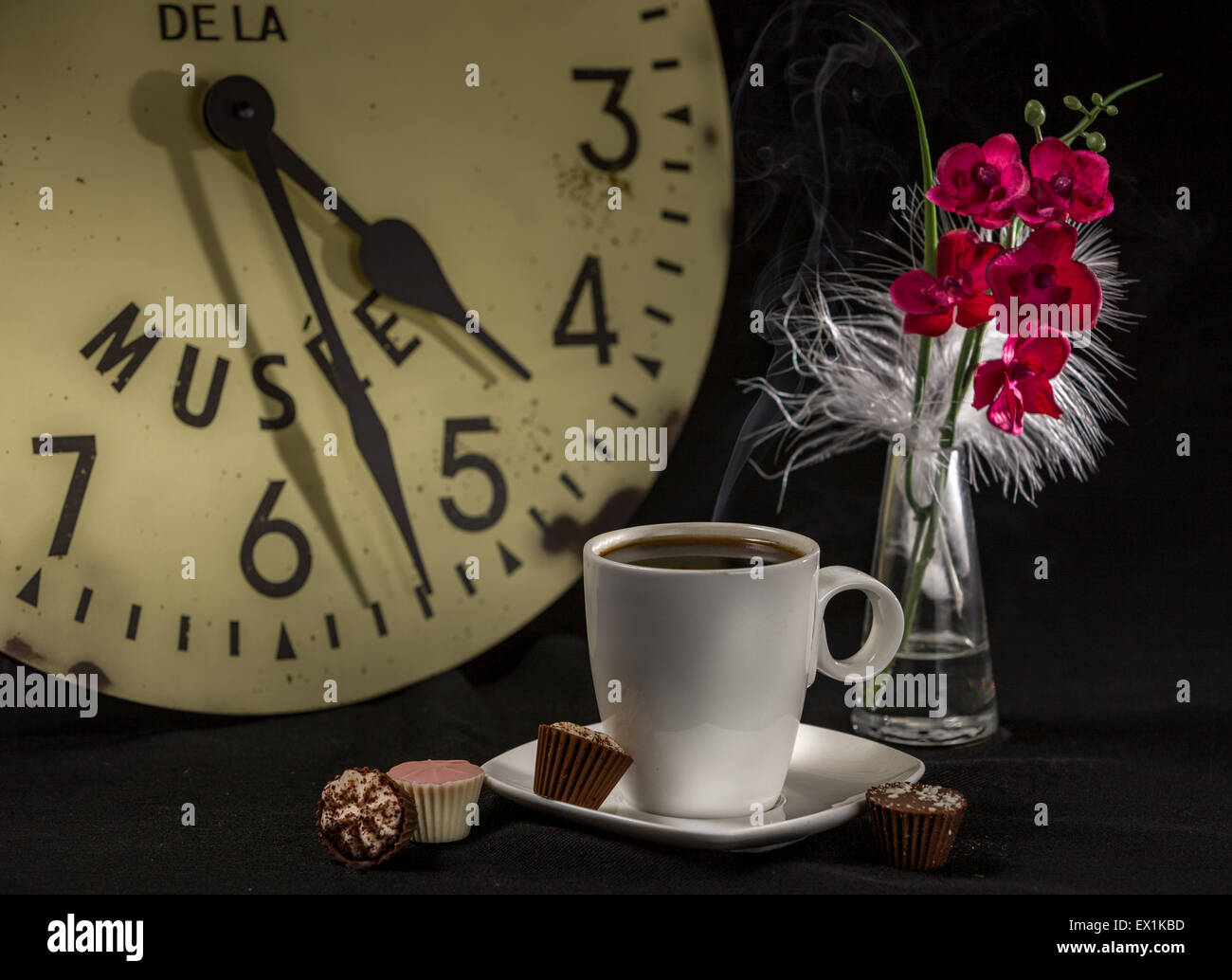 Stillleben mit einer Tasse Kaffee auf einem Tisch mit einer Uhr im Hintergrund;  eine Vase mit einer roten Blume und vier Pralinen Stockfoto