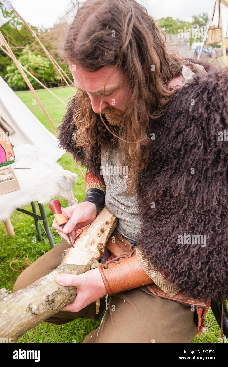 Ein Mann, gekleidet in der Kleidung der Eisenzeit schnitzt ein Gesicht in ein Stück Holz Stockfoto