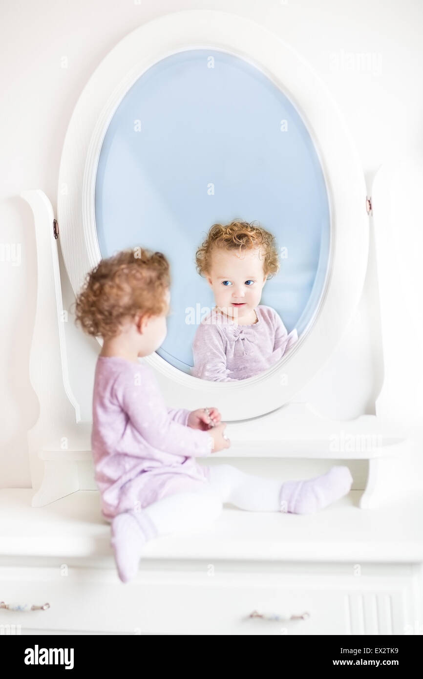 Niedliche kleine Kleinkind Mädchen mit dem lockigen Haar Blick auf ihr Spiegelbild in einem schönen weißen Spiegel Stockfoto