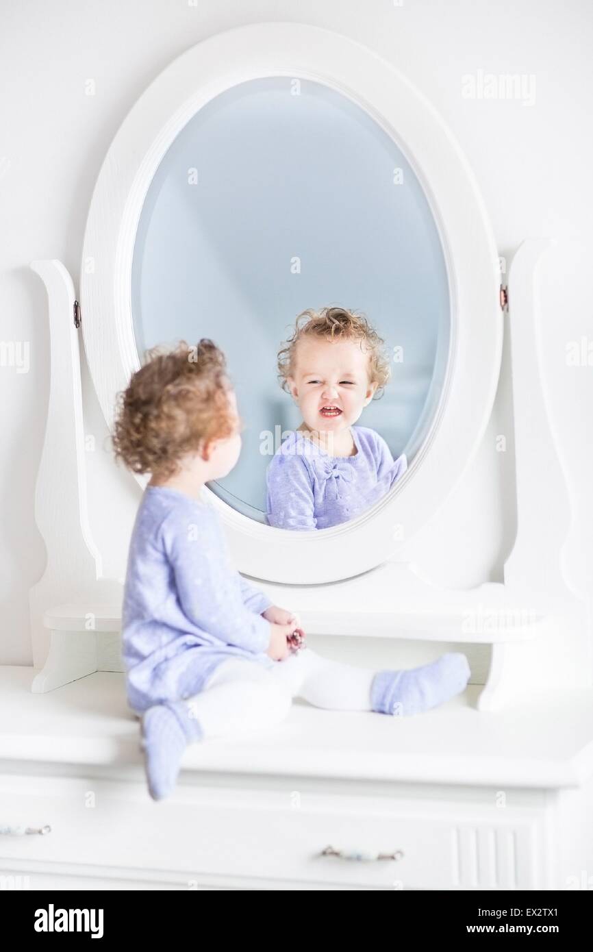 Entzückende lockige Kleinkind Mädchen lustige Grimassen beobachtet ihr Spiegelbild in einem schönen weißen Spiegel Stockfoto