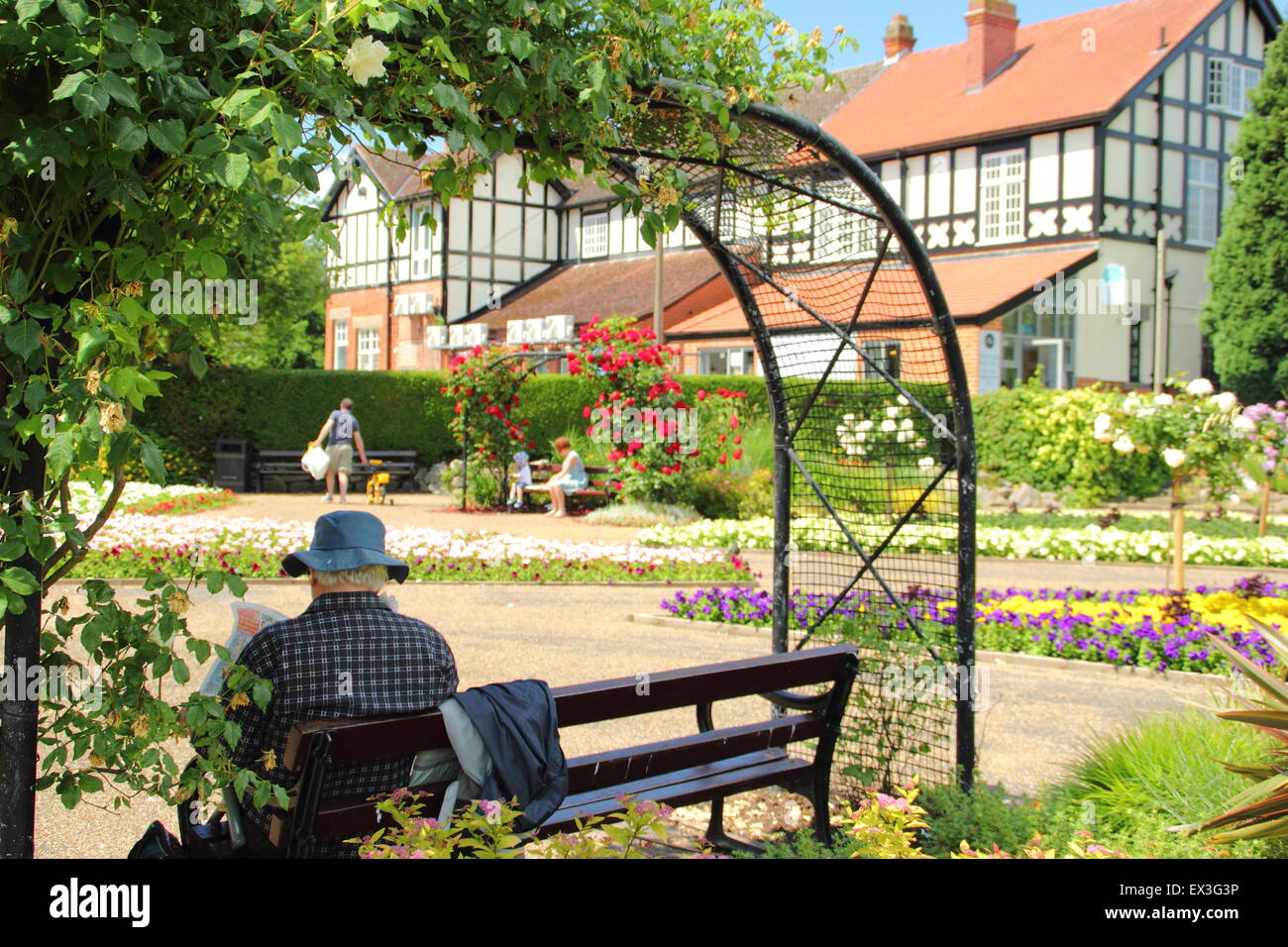 Ein Mann sitzt auf einer Bank unter einem rose Bogen in Hall Leys Park, einem traditionellen britischen Park in Matlock, Derbyshire, England UK Stockfoto