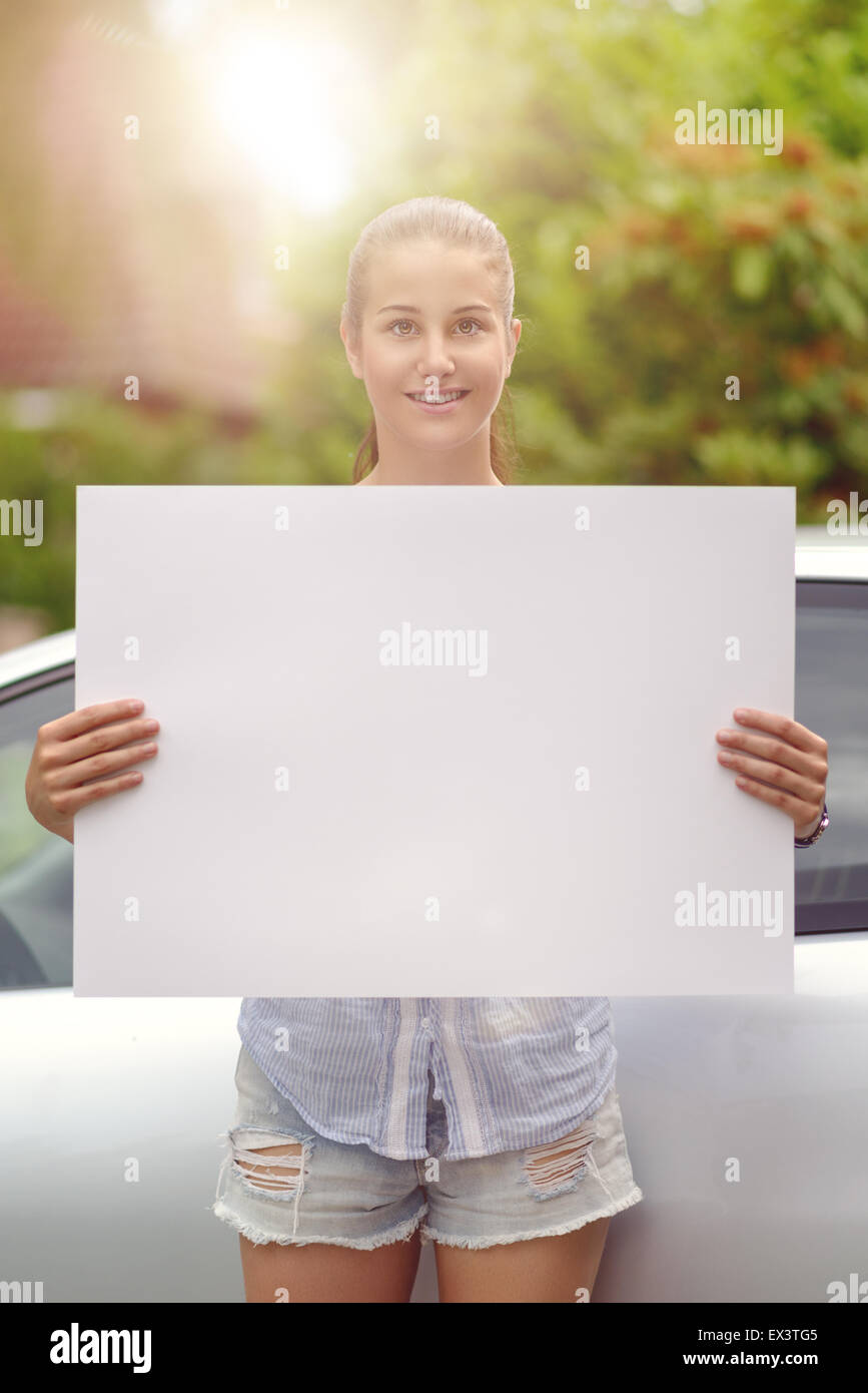 Hübsche junge Frau hält einen sauberen White Board mit textfreiraum und Lächeln in die Kamera vor ihrem Auto Stockfoto