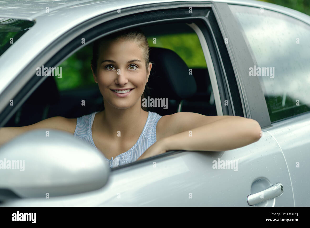 Attraktive junge Frau, die ein Auto aus ihrer offenen Seitenfenster in die Kamera schaut, mit einem warmen freundlichen Lächeln Lächeln Stockfoto