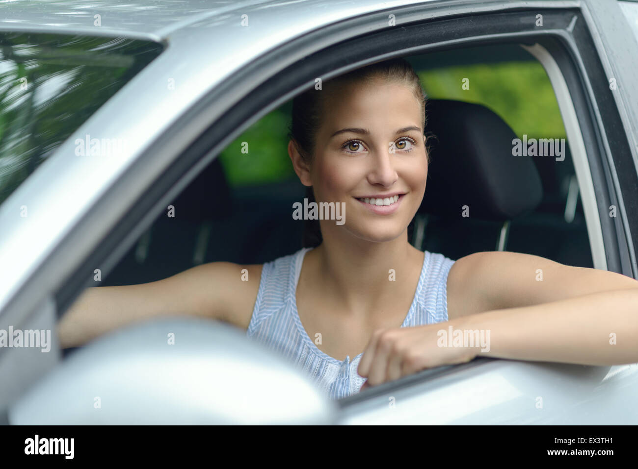 Attraktive junge Frau, die ein Auto aus ihrer offenen Seitenfenster in die Kamera schaut, mit einem warmen freundlichen Lächeln Lächeln Stockfoto