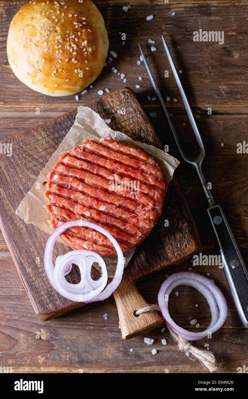 Rohe Schnitzel, in Scheiben geschnittene Zwiebel und frische Backen Brötchen für die Herstellung von hausgemachten Burger serviert auf kleinen Holzbrett mit Fleisch Gabel o Stockfoto