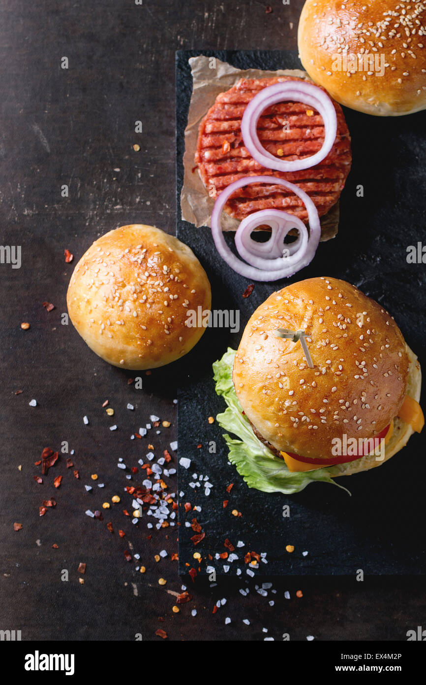 Frische hausgemachte Burger auf schwarzem Schiefer und rohe Schnitzel und in Scheiben geschnittene Zwiebel, serviert mit Meersalz und Pfeffer auf dunklem Hintergrund. Sele Stockfoto