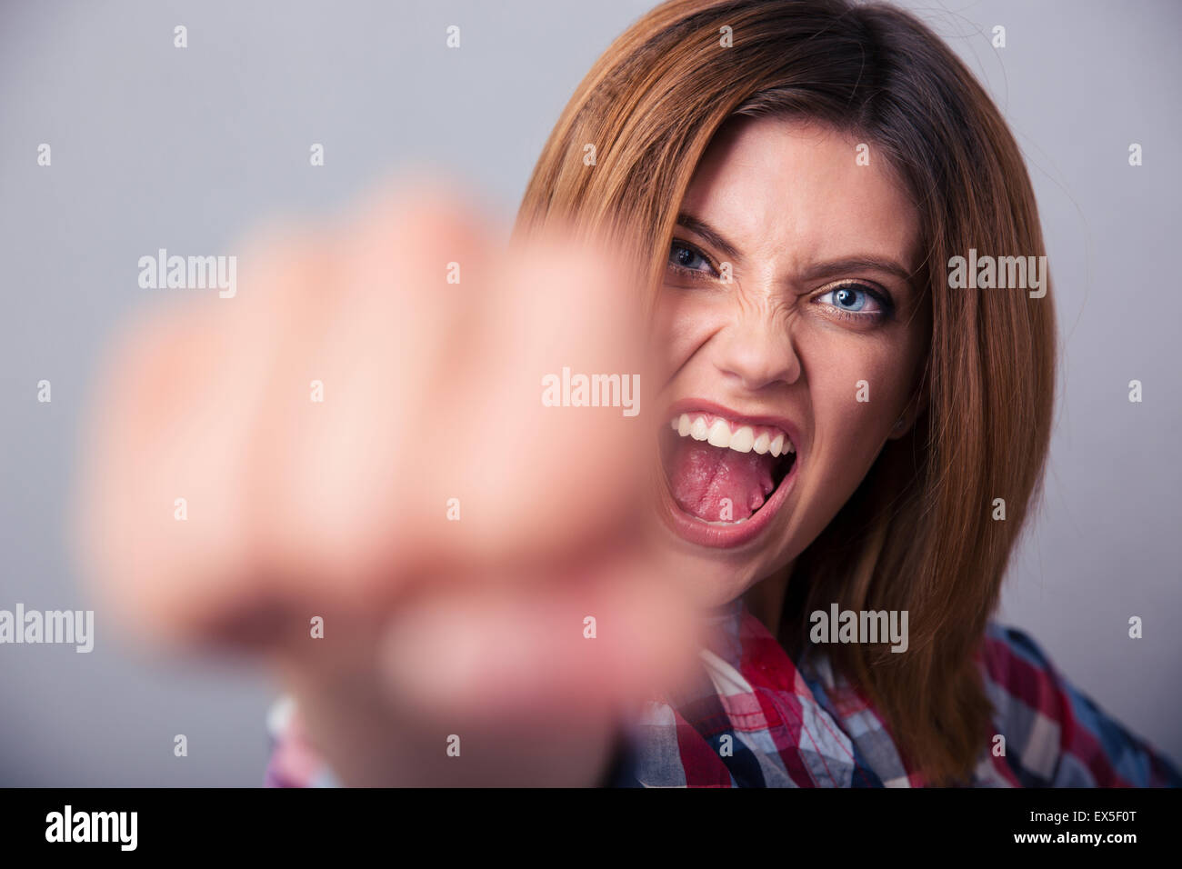 Böse Frau schlagen mit Faust vor der Kamera und schreien über grauen Hintergrund Stockfoto