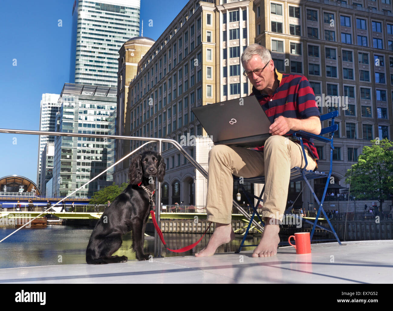 Er arbeitet von zu Hause aus, mit einem Hund, der draußen auf seinem Hausboot sitzt und an seinem Laptop Canary Wharf London E14 arbeitet Stockfoto