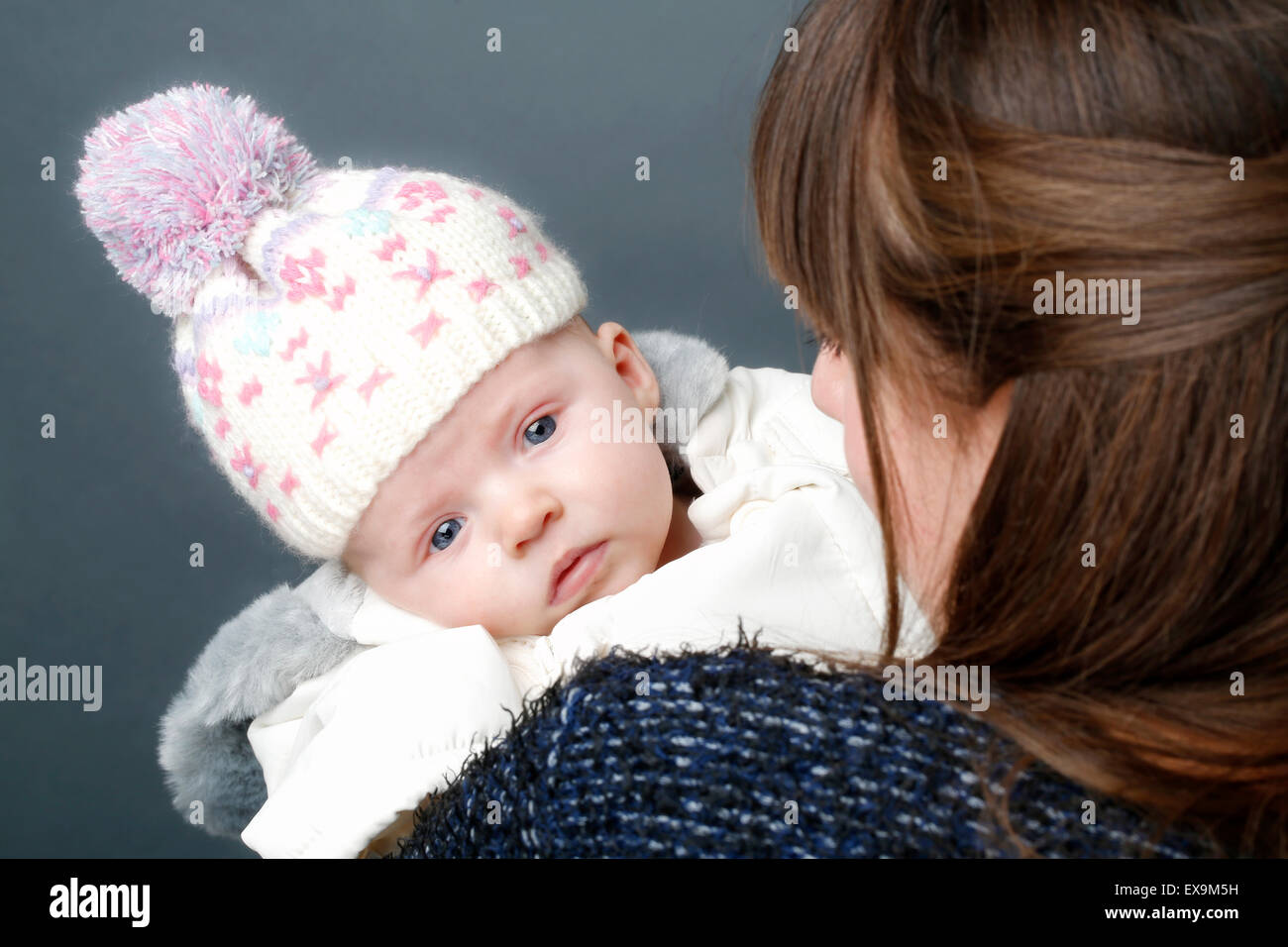 Ein kleines Baby, eingepackt für den Winter, mit einem warmen Hut und Mantel, das über der Schulter der Mutter auf die Kamera blickt Stockfoto