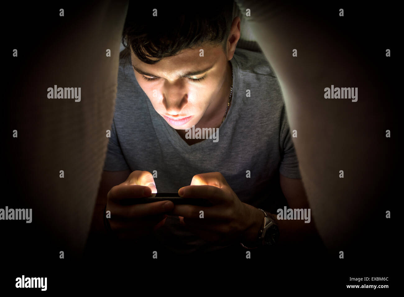 Teenager-Jungen versteckt sich unter einer Decke während der Benutzung eines Mobiltelefons für das Surfen im Internet oder privat zu chatten Stockfoto