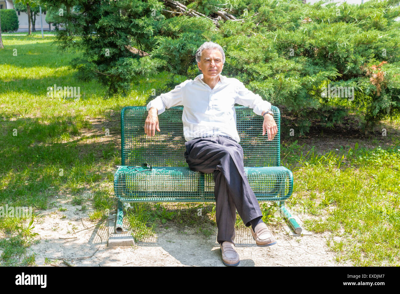 Hübscher Mann mittleren Alters mit Salz Pfeffer Haaren bekleidet mit weißem Hemd, blaue Hosen und Beige Mokassins ruht auf einer Bank im Stadtpark seine Arme geöffnet zu halten: er zeigt einen beruhigenden Blick Stockfoto