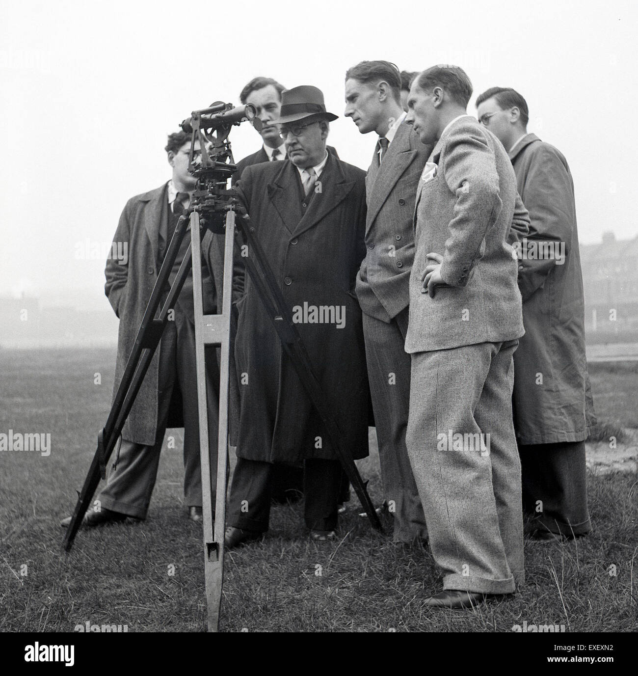 1950er Jahre, historisch, saß eine Gruppe männlicher Vermesser in Anzügen, Krawatten und Mänteln draußen auf einem Feld mit einem Theodoliten auf einem großen Stativ. Ein präzises optisches Instrument, ein Theodolit, wird für topographische Vermessungen und für den Bau von Gebäuden und Infrastrukturen eingesetzt. Stockfoto