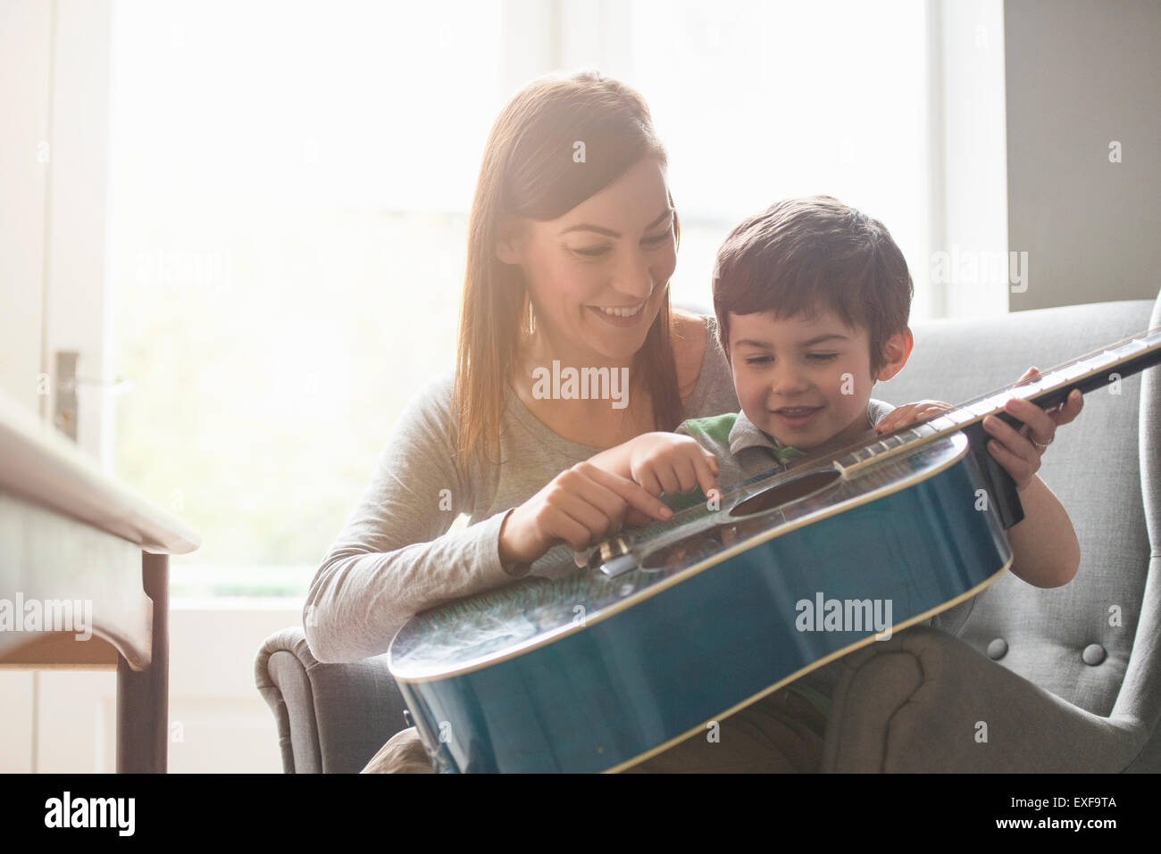 Junge auf Mutters Schoß im sonnendurchfluteten Zimmer sitzen und lernen, Gitarre zu spielen Stockfoto