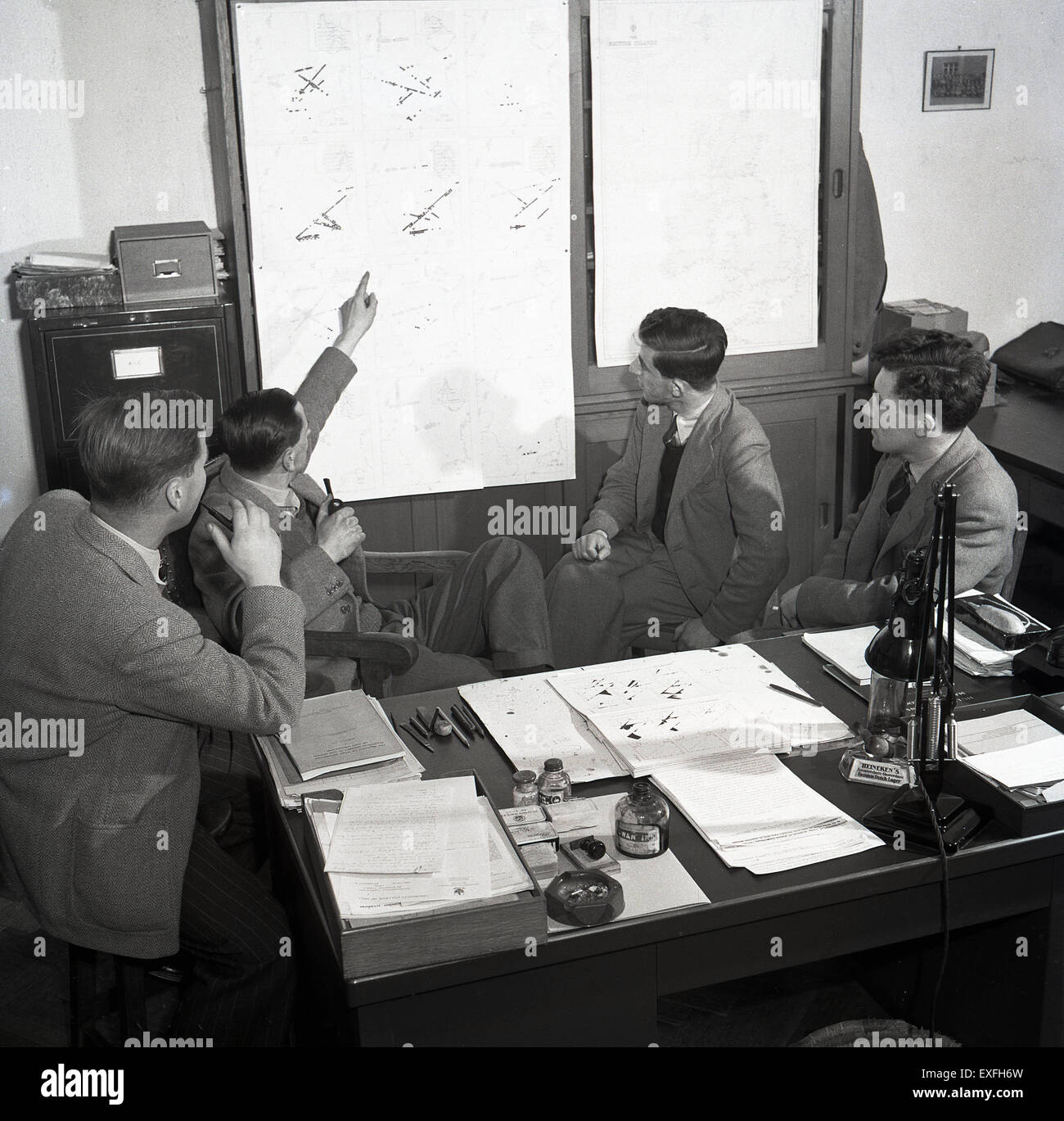 1950er historische Ingenieurstudenten werden im Büro eines Professors in England unterrichtet. Drei junge Männer sitzen neben ihrem Lehrer an seinem Schreibtisch, der auf Informationen auf einem Blatt zeigt. Stockfoto