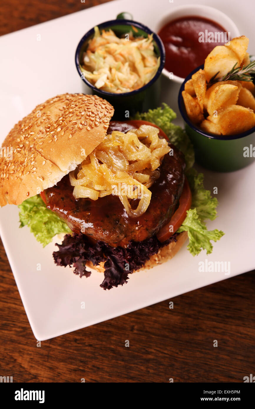 Köstlich saftige Rindfleisch-Burger mit karamellisierten Zwiebeln und Salat serviert mit Kartoffelecken auf einem weißen Teller auf einem Holztisch. Stockfoto