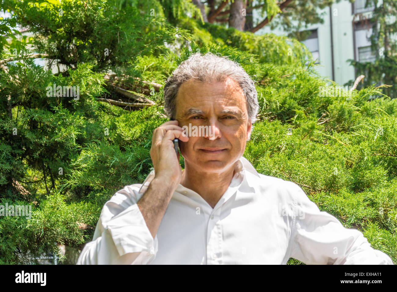 Hübscher Mann mittleren Alters mit Salz Pfeffer Haar gekleidet mit weißen Hemd ist telefonieren mit Handy im Stadtpark: er zeigt einen beruhigenden Blick Stockfoto