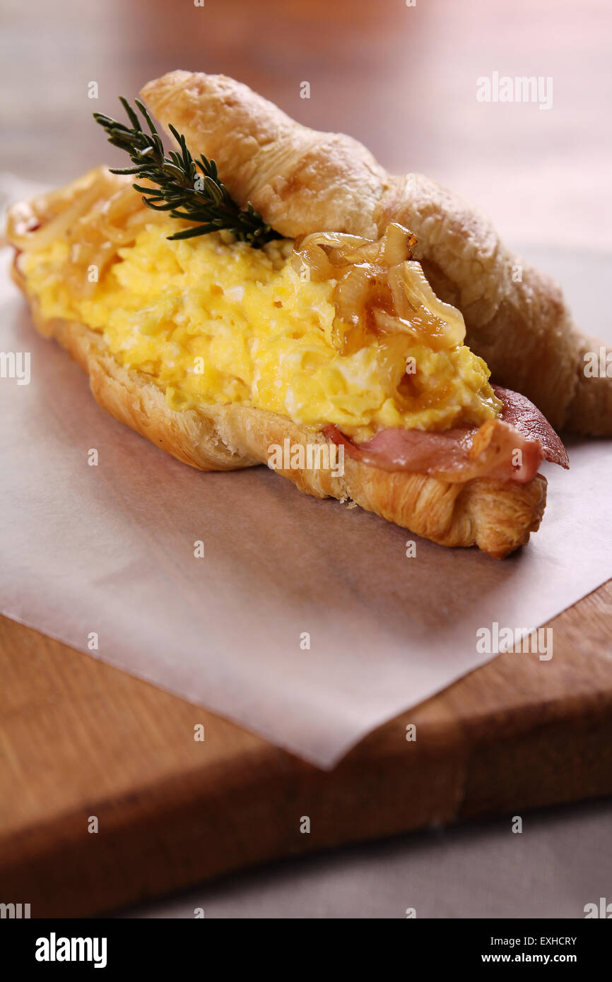 Auf eine Holzplatte auf einem Holztisch serviert leckere Croissant mit Speck, Rührei Ei karamelisiert Zwiebeln und Rosmarin. Stockfoto