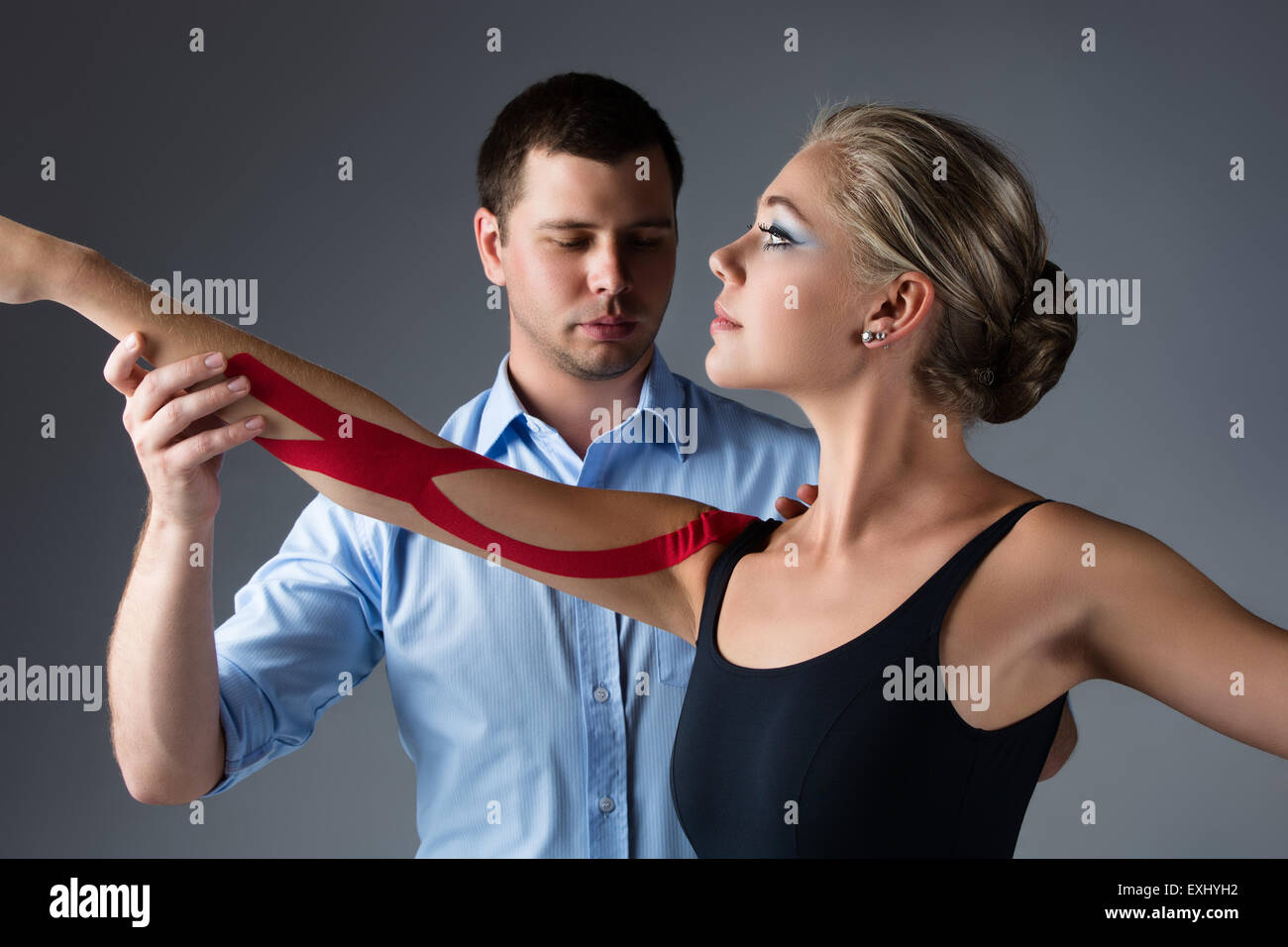 Schöne weibliche Ballett-Tänzerin und ihr Physiotherapeut auf grauem Hintergrund. Ballerina trägt einen schwarzen Anzug und hat rote st Stockfoto