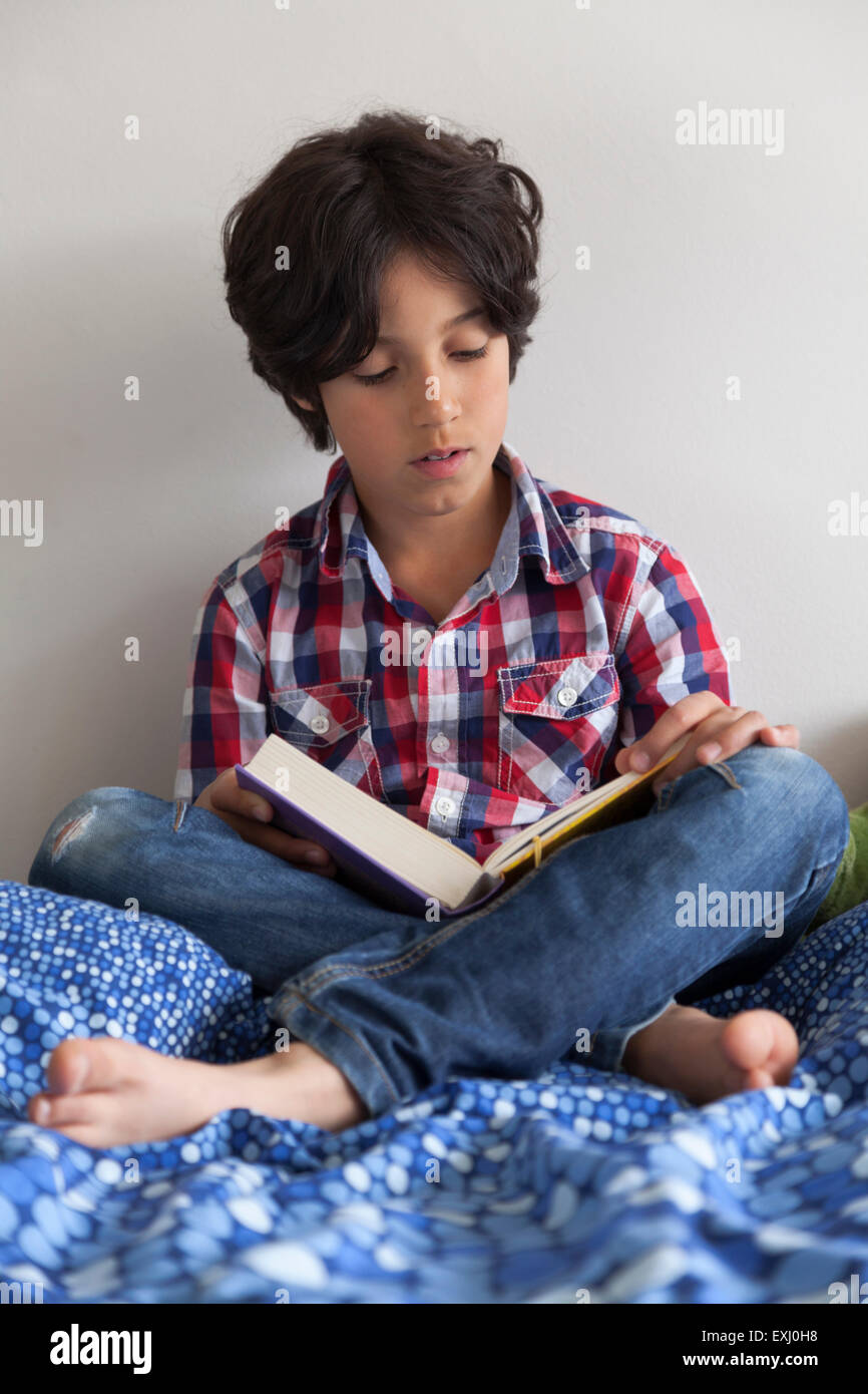 Zehn Jahre alte Junge liest ein Buch auf seinem Bett sitzend Stockfoto