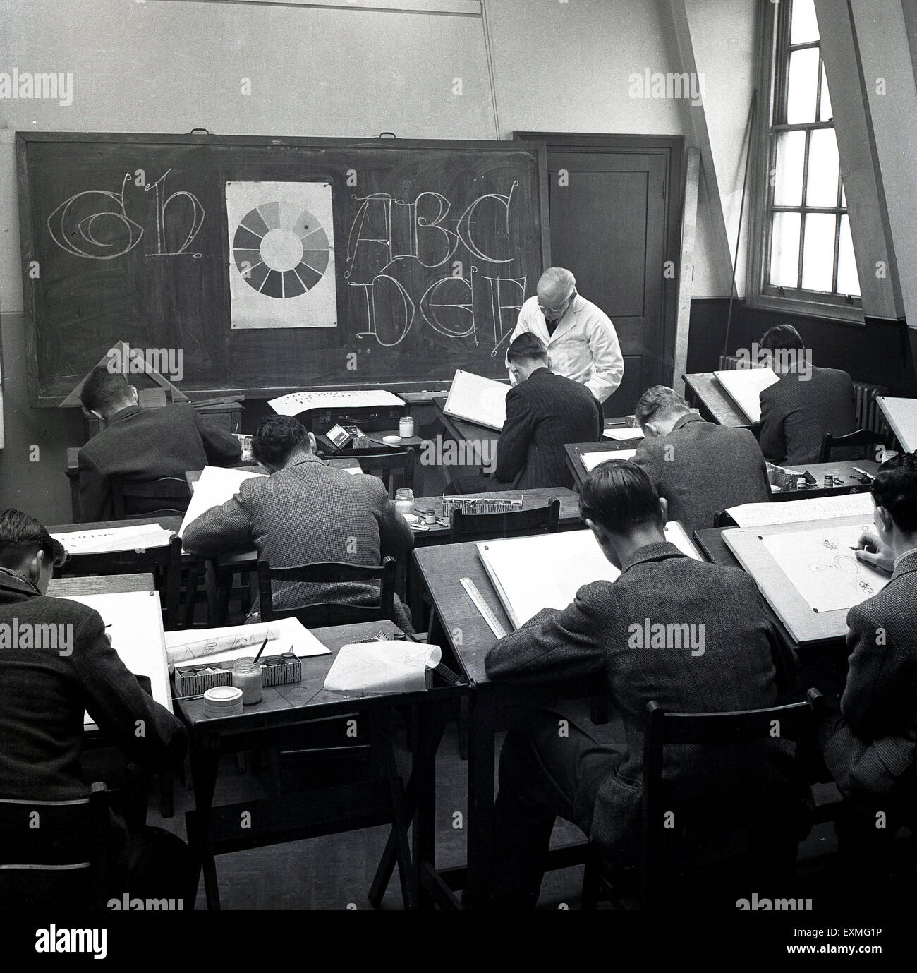 1950er Jahre, historisches Bild zeigt eine Gruppe von männlichen Studenten sitzen an traditionellen einzelnen hölzernen Schreibtischen in einem Klassenzimmer unterrichtet Grafikdesign von einem weißen überzogenen männlichen Lehrer, der einem Schüler an der Tafel hilft. Stockfoto