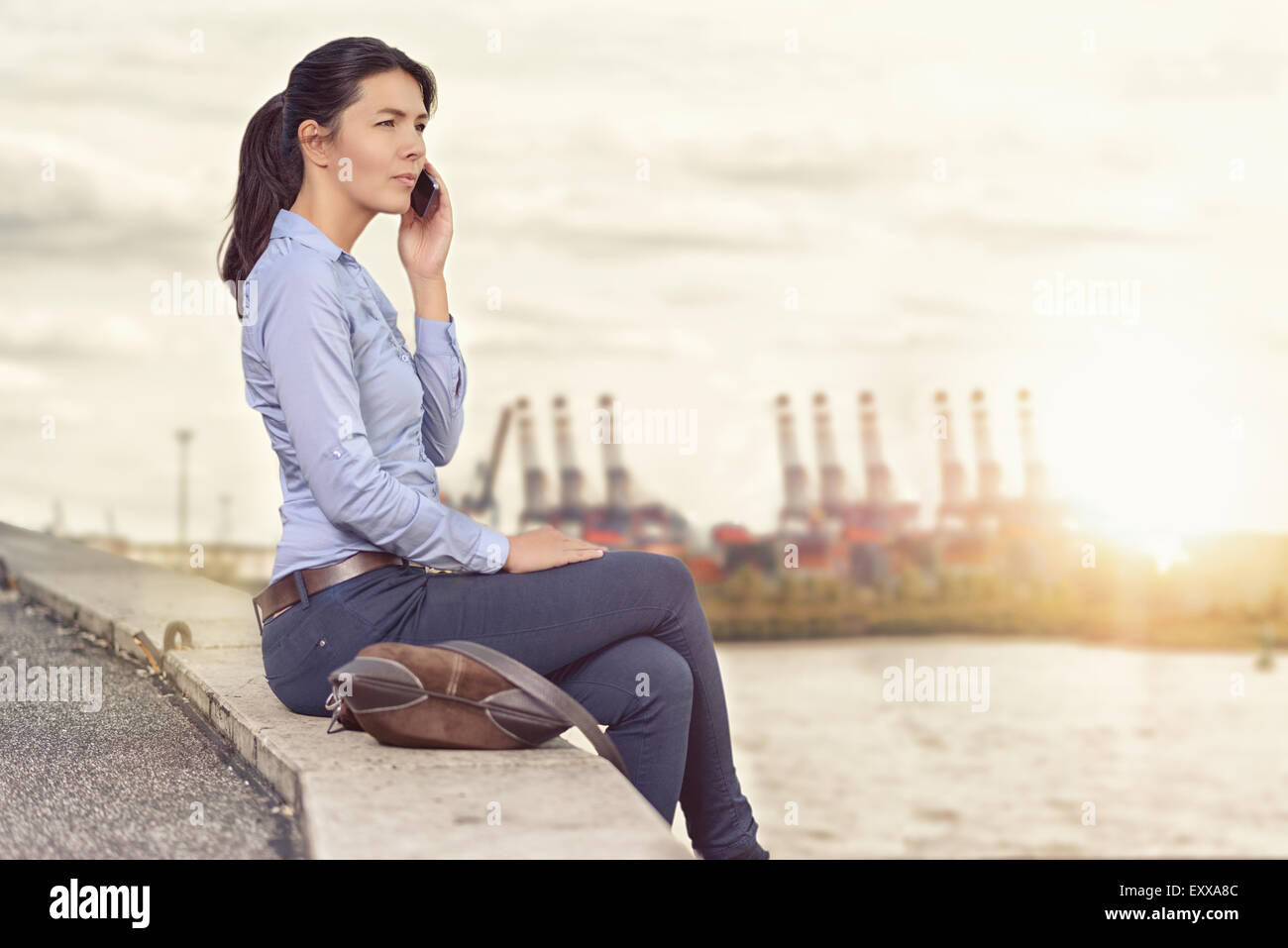 Attraktive stilvolle Frau, die auf ihrem Handy zu sprechen, als sie auf eine Wand mit Blick auf einen Fluss und entfernten Stadt sitzt Seitenansicht mit Stockfoto