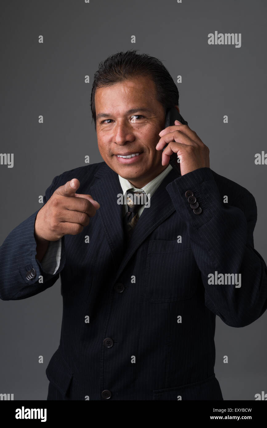 Hispanic Geschäftsmann hält eine Zelle Telefon, Blick in die Kamera und lächelnd zeigte mit einen Finger. Stockfoto